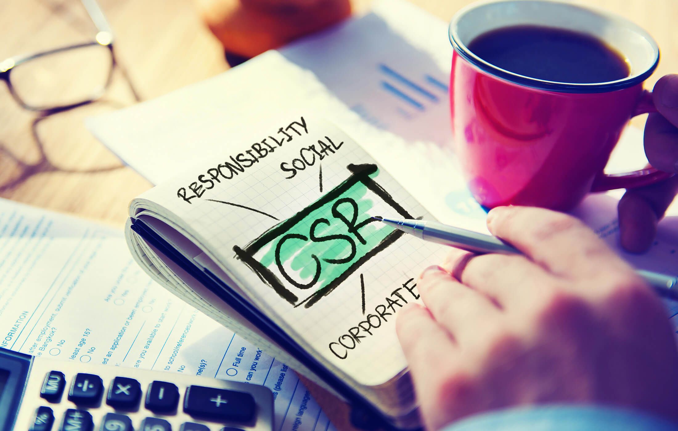 Kockás jegyzetfüzet lapján bekeretezett, zölddel kiemelt CSR felirat, körülötte a RESPONSIBILITY, SOCIAL és CORPORATE szavak; az asztalon toll, kávéscsésze, szemüveg és számológép